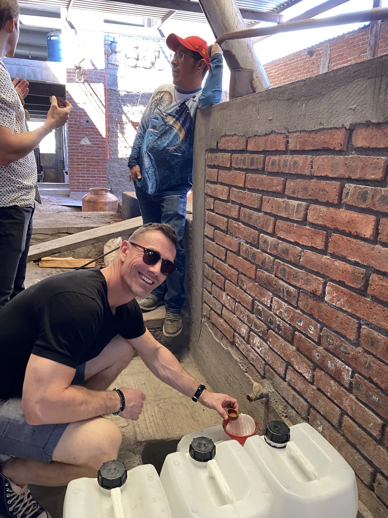 A man in sunglasses crouches to fill containers from the still at a brick palenque while the team looks on