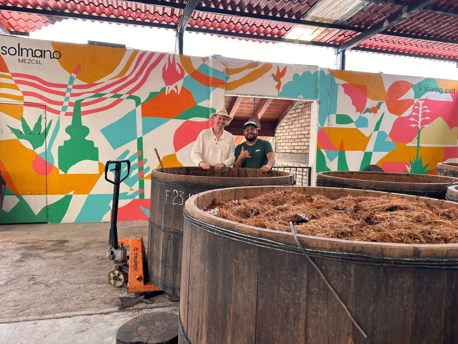 Two men stand behind wooden fermentation vats of agave fibre at the Solmano palenque, in front of a colourful Solmano mural