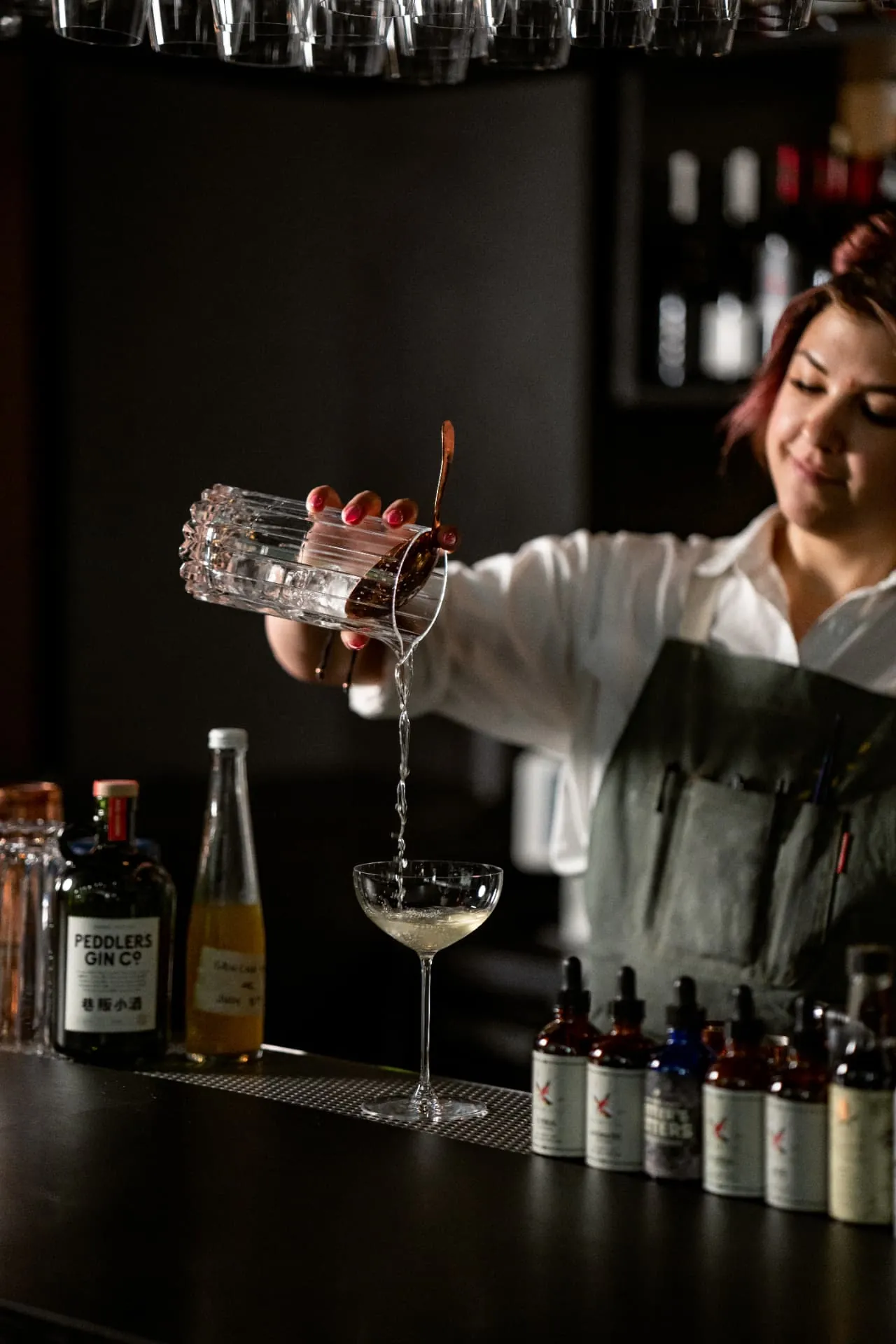 A bartender strains a stirred gin cocktail into a coupe, a bottle of Peddlers Gin on the bar