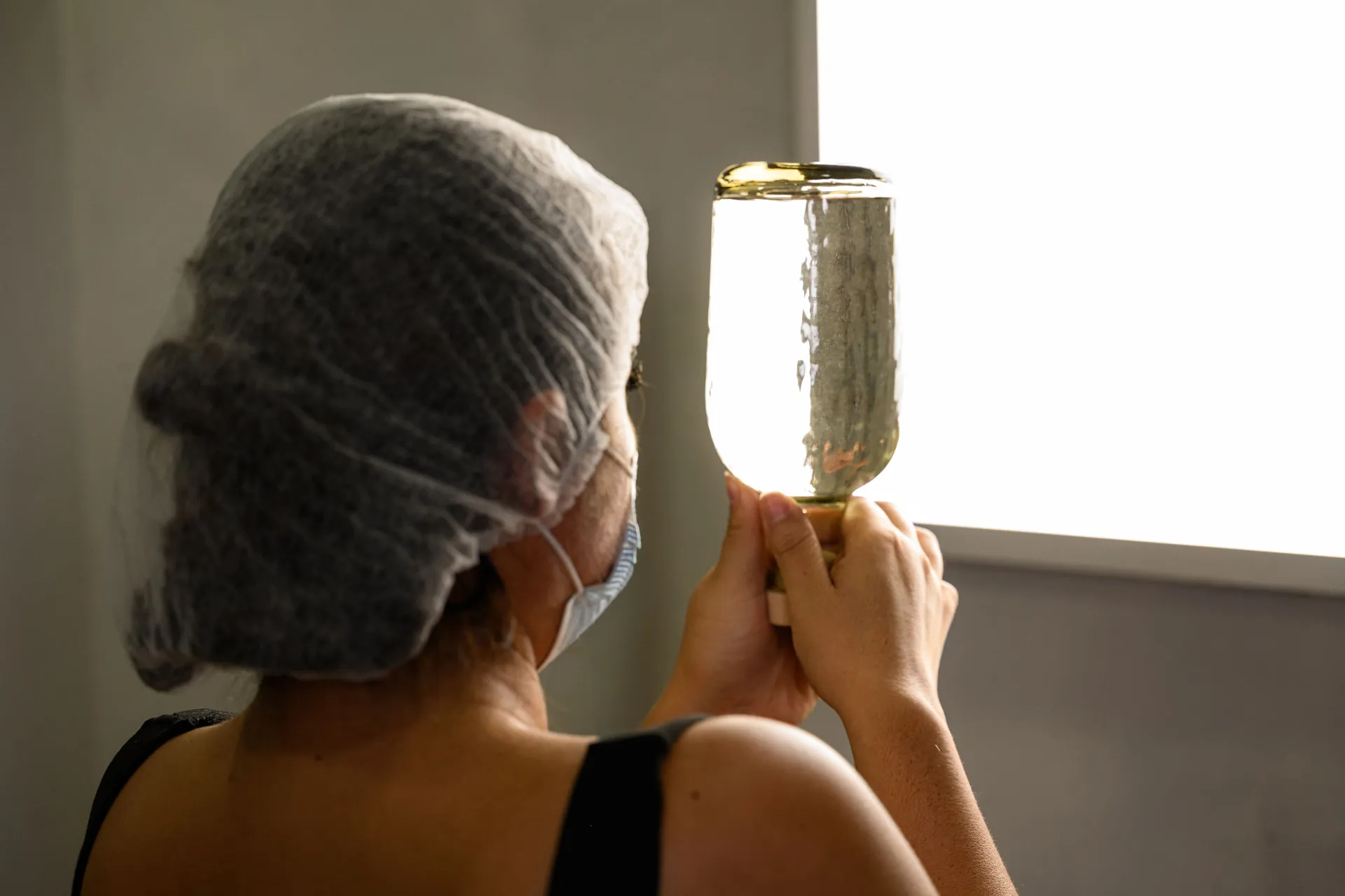 A worker in a hairnet holds a bottle of NYC Tequila up to a light panel to inspect the liquid