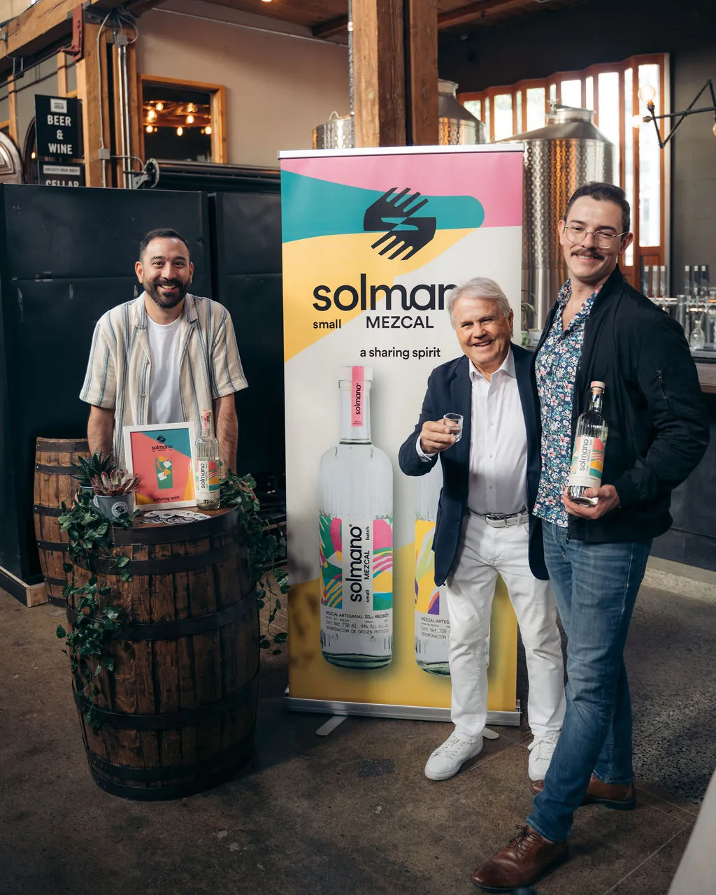 Three people smile beside a Solmano Mezcal banner at a launch event, one raising a copita